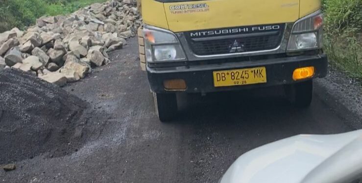 Potret kendaraan proyek yang berpapasan dengan kendaraan warga di jalan yang bahu jalannya ditumpuki oleh material proyek.